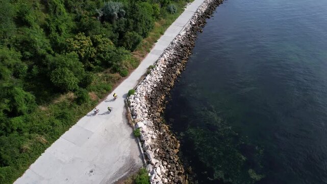 Aerial view of cyclists riding along a concrete promenade next to a rocky breakwater and lush green trees in Balchik, Dobrich Province, Bulgaria.