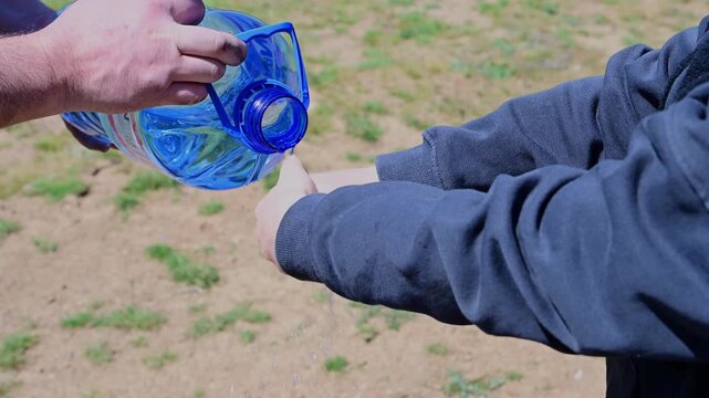 one person pours fresh water from a large blue plastic bottle over the cupped hands of another person, providing a simple way to wash hands in a rural or natural setting.