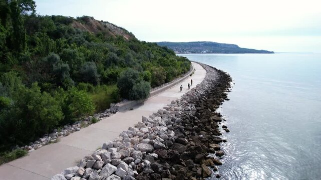 Aerial view of people cycling along a coastal promenade with a rocky breakwater and lush green hills under a bright sky in Balchik, Dobrich Province, Bulgaria.