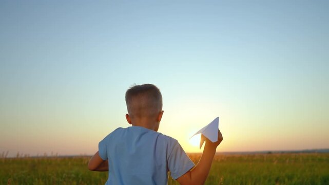 Boy runs with paper airplane in field at sunset. Child plays with paper plane outdoors. Kid flying aircraft in meadow. Boy running with airplane. Child plays with paper glider at golden hour.