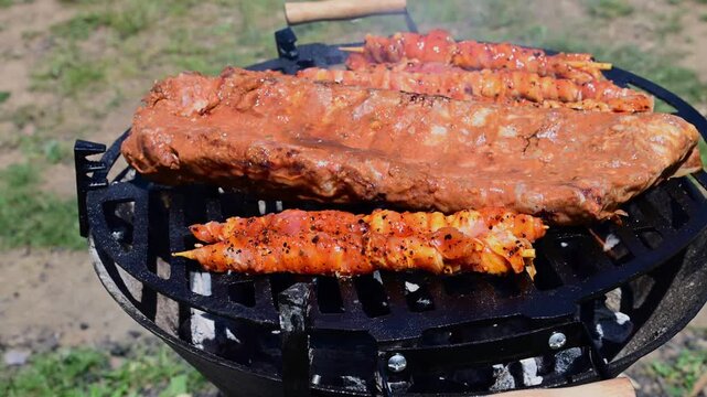 marinated pork ribs and small meat skewers sizzling on a circular charcoal grill during an outdoor barbecue.