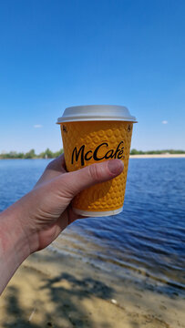 Kyiv, Ukraine - May 6, 2026: Yellow paper cup of McCafe coffee in hand on the background of the river and beach. Female hand holds a disposable cup with coffee from McDonald's. Drink.