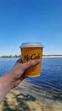 Kyiv, Ukraine - May 6, 2026: Yellow paper cup of McCafe coffee in hand on the background of the river and beach. Female hand holds a disposable cup with coffee from McDonald's. Drink.