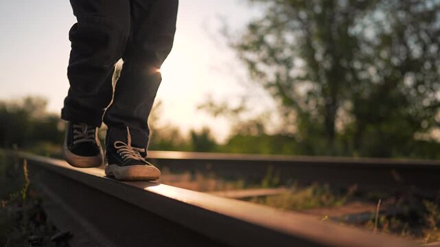 Child is walking on railroad track at sunset. Boy balances on rail in sneakers during summer evening. Legs step on train tracks outdoors. Kid walks away in nature with golden sun light in background.