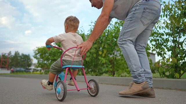 Caring father pushes little son on tricycle along path in summer park. Happy boy learns to ride bike with dad helping outdoors. Parent walks beside child on tricycle. Family enjoys leisure time.
