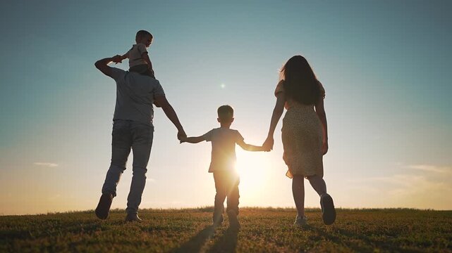 Happy family is walking in green field at sunset. Father carries son on shoulders while walking with mother. Parents hold hands with child. Family walks away in nature park during summer evening.