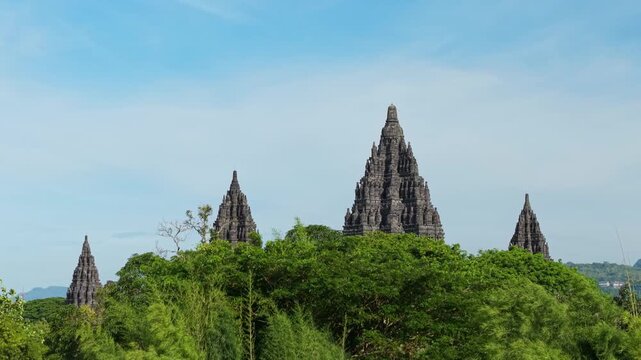 Aerial view of the ancient Prambanan Temple spire peaking through dense tropical foliage under a clear blue sky in Kecamatan Prambanan, Daerah Istimewa Yogya, Indonesia.