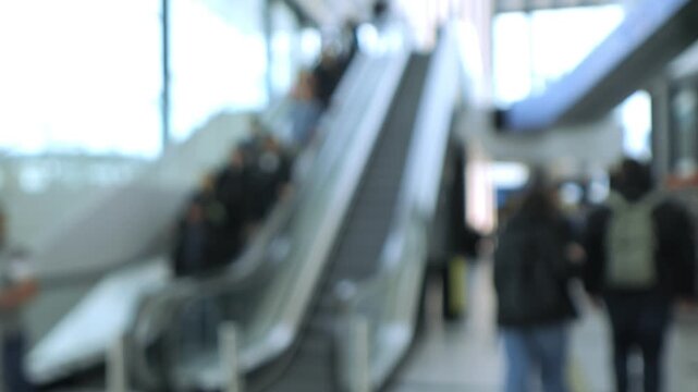 Blurred footage shows people moving on escalators in a busy indoor space. Figures appear out of focus as they travel up and down. Background suggests a modern transit area or shopping mall