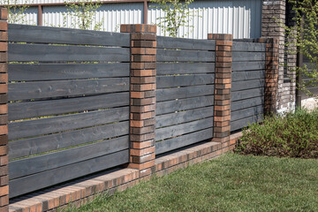 Fototapeta premium Dark horizontal wooden fence planks with red brick pillars and base near green lawn, residential property boundary and perimeter wall