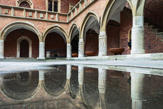 Courtyard of Collegium Maius in Krakow with late Gothic arcades and cloisters on a cloudy day after rain, with architectural reflections visible in puddles