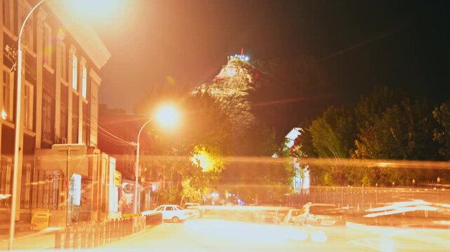 Timelapse Sulayman Mountain or Sulaiman-Too at night in Osh, Kyrgyzstan. Illuminated sacred peak and flag, overlooking glowing city street with warm streetlights and parked cars.