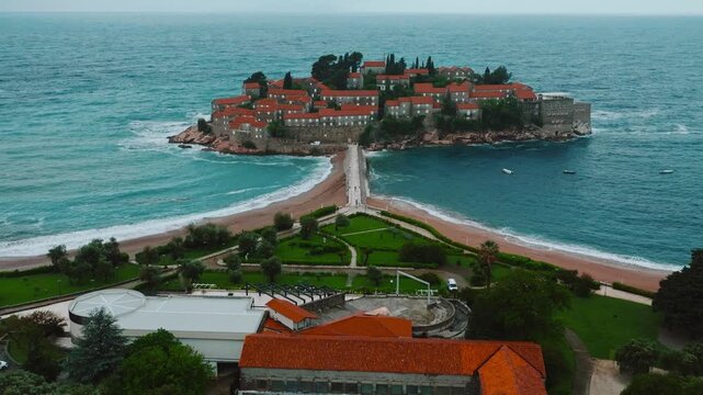 Cinematic aerial drone footage of Sveti Stefan featuring the iconic island village connected to the mainland by a narrow causeway, surrounded by turquoise Adriatic waters