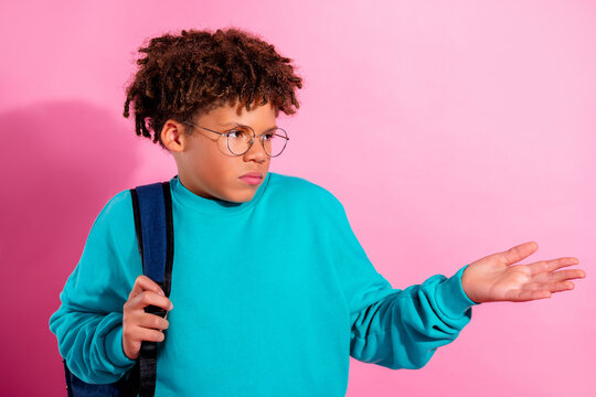Teen boy shrugging with backpack and glasses against pink background showing confused, indifferent expression in turquoise sweater