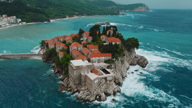 Cinematic aerial drone footage of Sveti Stefan featuring the iconic island village connected to the mainland by a narrow causeway, surrounded by turquoise Adriatic waters