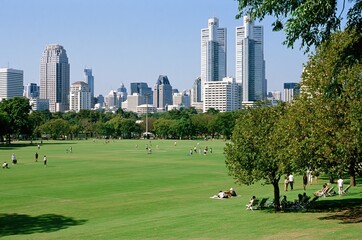 Obraz premium Sprawling green park with people relaxing and playing near a modern city skyline under a clear blue sky