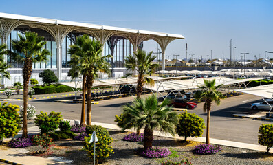 Naklejka premium The modern exterior design and landscaping of Prince Mohammad bin Abdulaziz International Airport in Madinah, Saudi Arabia, showing the terminal and parking area.