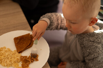 Left handed little child eating pancakes with a fork sitting at the dinner table. Selective focus