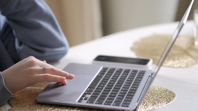 Hand taps laptop touchpad. White table with gold textured placemat. Smartphone lies beside laptop. Person wears gray sleeve and ring. Soft natural light from side