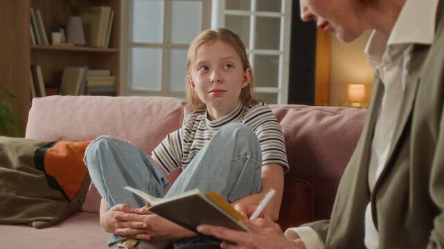 Rack focus shot of preteen girl answering therapists questions sitting on couch in living room while specialist note taking during psychological assessment session