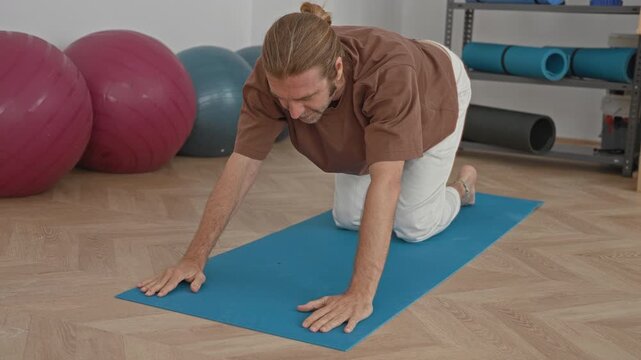 Man on yoga mat performing gentle backbend with hands pressed to wooden floor in studio; calm concentration.