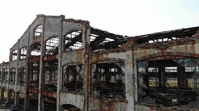 A weathered industrial building, showing decay with broken windows, arches, and structural damage