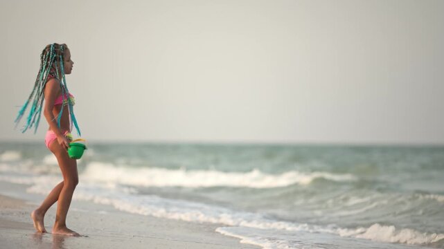 A girl with african braids in a summer costume plays on the beach with shells near the sea with waves under sunny sunset