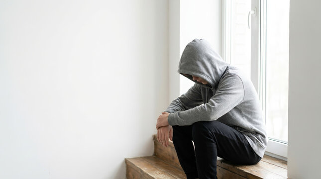 Adolescent hiding behind hoodie sitting on stairs in a bright room with window light