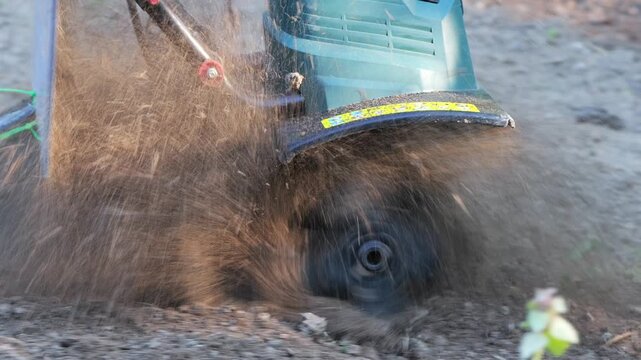 Slow motion close up video shows electric garden cultivator throwing soil and dust while rotating blades loosen dry ground in outdoor gardening environment 2026