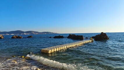 Mountains and a port are visible in the hot haze on the seashore. A plastic pier is installed in transparent water near the shore and large stones are lying. A ripple and buoys on the water. Sunny © Balser