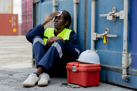 A tired female worker in safety gear sits on the ground wiping sweat from her forehead at a shipping container yard.