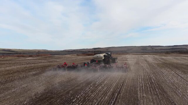 Cinematic aerial footage of a modern tractor pulling an air seeder across rolling prairie farmland during spring planting season