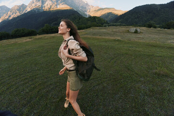 Naklejka premium A woman hikes outdoors with a backpack through a grassy valley, with mountains in the distance, conveying adventure, exploration, and a sense of freedom.