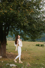Naklejka premium Woman lifestyle portrait outdoors, a figure stands under a large tree in a sunlit field, wearing white outfit, relaxed pose, serene mood and natural setting.