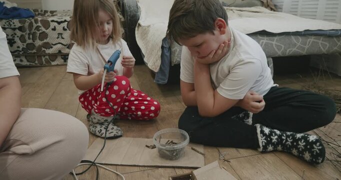 A creative family spends time together. A little girl uses a glue gun to make a craft while her brother watches, and her mother helps them from the floor at home.