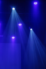 Naklejka premium Blue and purple stage lights cast bright beams through haze from a dark ceiling rig, creating a dramatic performance background with copy space