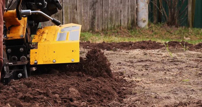 Close-up of a farmer using a yellow walk-behind tractor to plow and cultivate the soil, preparing the land for planting in a backyard garden during the agricultural season.
