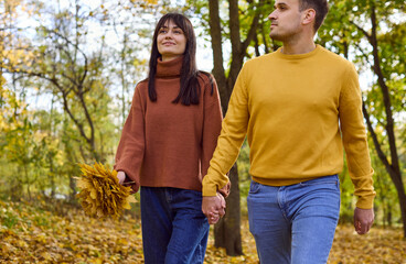 Fototapeta premium Couple walking together in autumn park, sharing love. They stroll a path of fallen leaves, sharing happiness, romance, and a caring relationship. Symbol of romance and seasonal happiness.