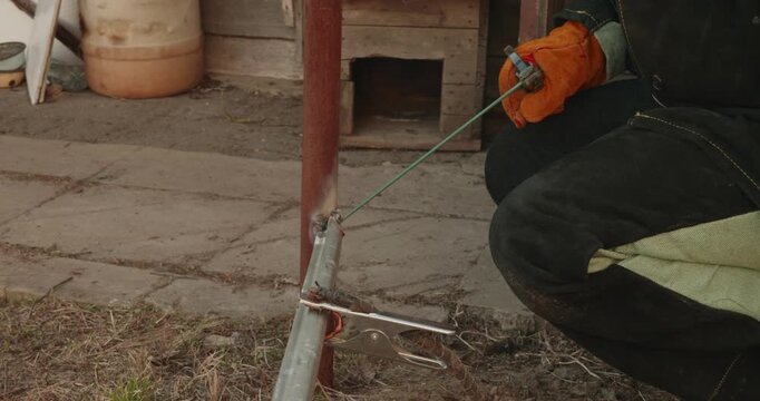 Skillful welder wearing protective gloves using an arc welding machine to join metal components, creating bright sparks and smoke while repairing a fence in a backyard construction site.