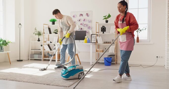 Woman and man cleaning apartment, vacuuming and mopping. Both wear aprons and gloves, tidying a bright interior with vacuum, mop and blue bucket. Teamwork keeps household chores on track.