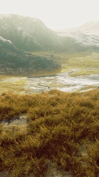 A grassy area with patches of water is seen. Mountains rise in the background under a bright sky. The scene shows a natural environment with open space and distant hills.