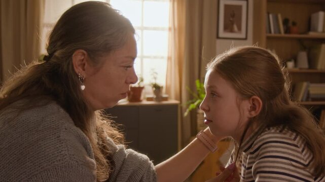 Side view of middle aged woman gently tucking daughters hair behind eras then kissing young girl on forehead while sharing heartfelt moment together in sunlit living room