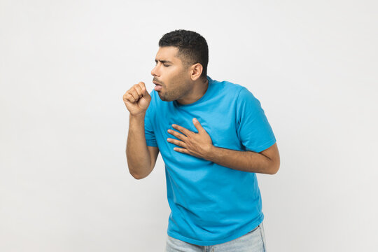 Portrait of sick unhealthy ill unshaven man wearing blue T- shirt standing coughing, having grippe or flu symptoms, feels unwell. Indoor studio shot isolated on gray background.