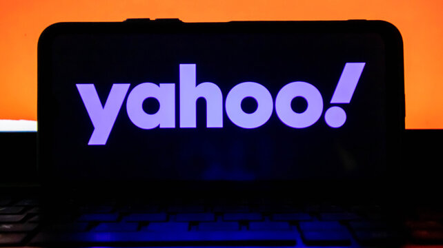 Closeup of the yahoo logo displayed on a smartphone screen with an orange background and a keyboard in the foreground