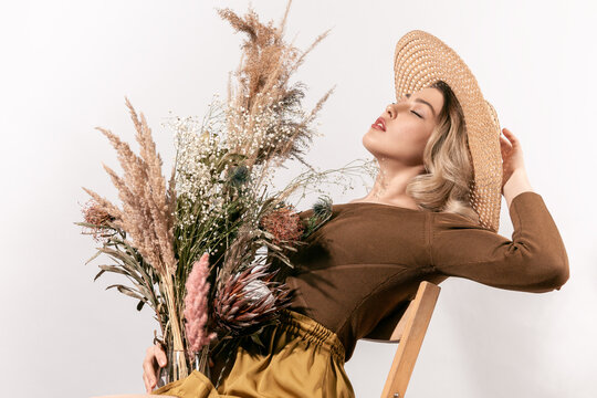 Young woman in hat leaning back on wooden chair holding bouquet of dried flowers with closed eyes and relaxed face expression isolated on white background