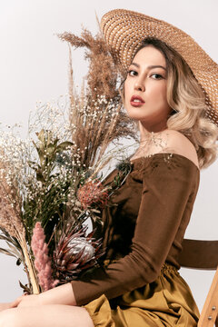 Elegant female model in hat holding dried flowers vase sitting on chair looking at camera with open mouth and expressive face showing summer style and autumn floral mood isolated on gray background