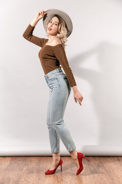 Fashionable young woman posing in red high heels and blue jeans wearing brown off-shoulder sweater holding grey hat on head standing in profile near white wall in studio