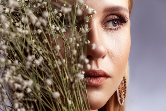 Young woman face with professional makeup partially covered by dried flowers branches isolated over gray background