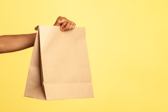 Side view portrait of bearded deliveryman wearing red visor cap and blue overalls, giving paper parcel to client, fast delivery service. Indoor studio shot isolated on yellow background.