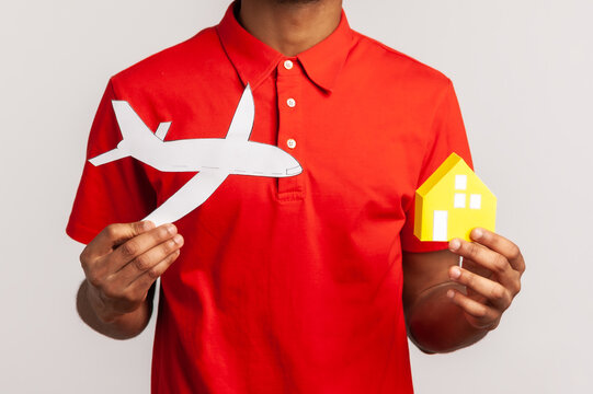 Smiling man with dreadlocks in red casual style T-shirt, holding paper plane and house, planning to move abroad, buying apartments in another country. Indoor studio shot isolated on gray background.