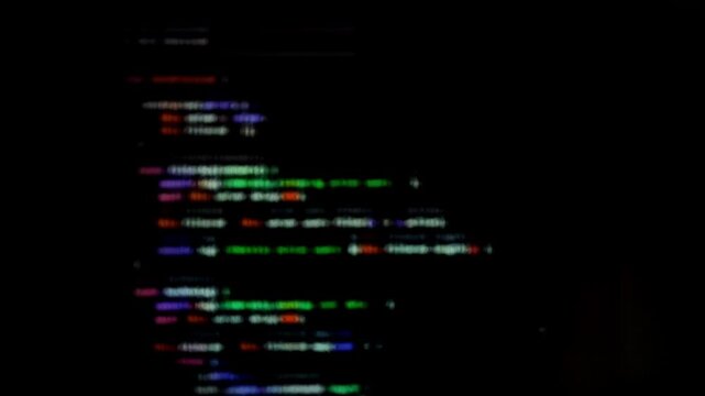 Colorful blurred code moves on dark screen creating abstract programming background. Defocused script lines form dynamic digital texture. Technology concept reflects modern coding